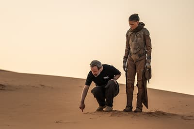 Behind-the-scenes photo of Denis Villeneuve crouching on a dune beside Zendaya in costume as Chani