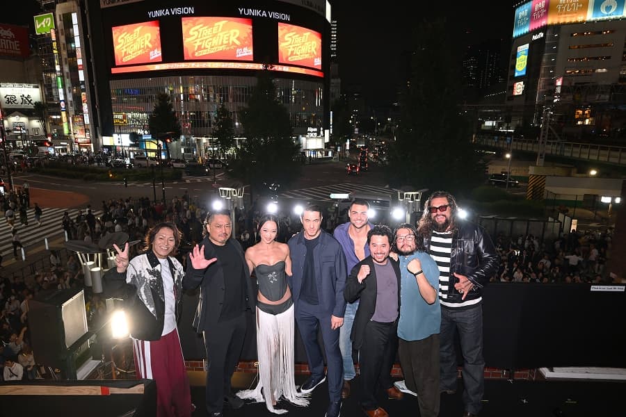 Cast and director on a balcony in Shinjuku at night, with Street Fighter logos on the Unicus Vision screens behind